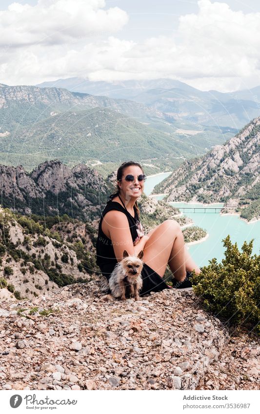 Anonymous female tourist with dog sitting on hill and enjoying landscape woman summer trip scenery nature vacation rest pet sunglasses black picturesque journey