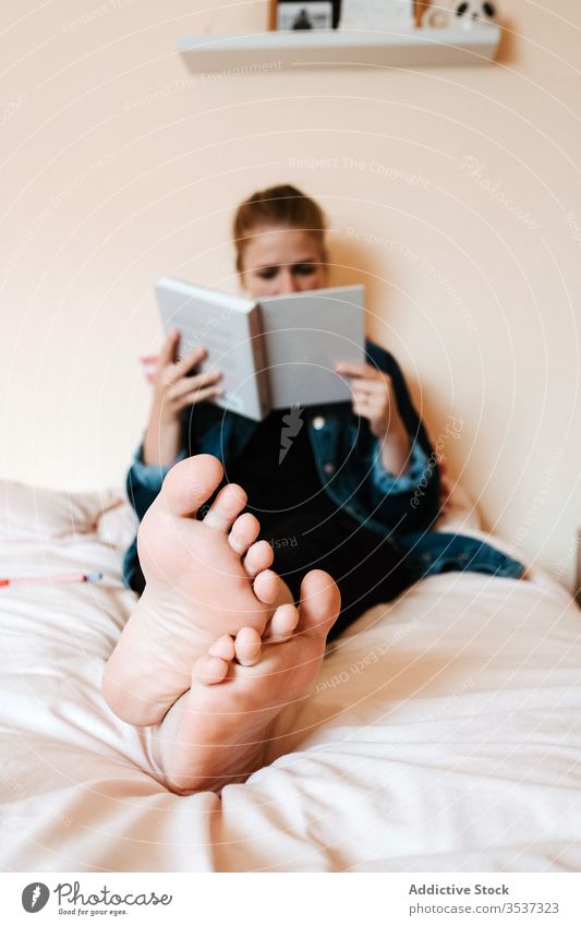 Pensive young woman reading book in light bedroom home barefoot bookworm alone relax textbook student rest chill recreation entertain calm lounge story female