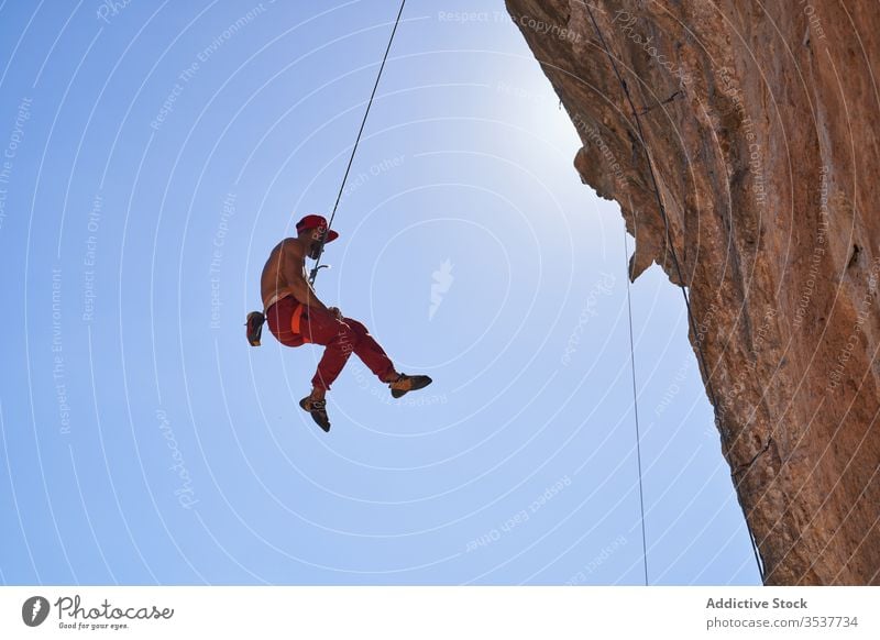 Strong male climber hanging on rope near rocky slope of mountain on sunny day man mountaineer strong sport secure equipment nature adventure activity cliff