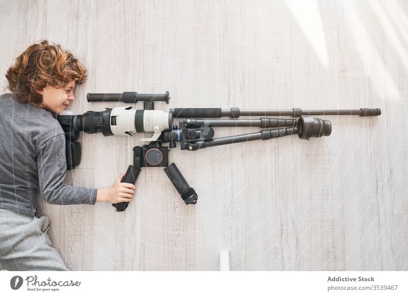 Young photographer playing with accessories for professional camera like with rifle while lying on floor boy photo camera sniper game pretend concept shooter