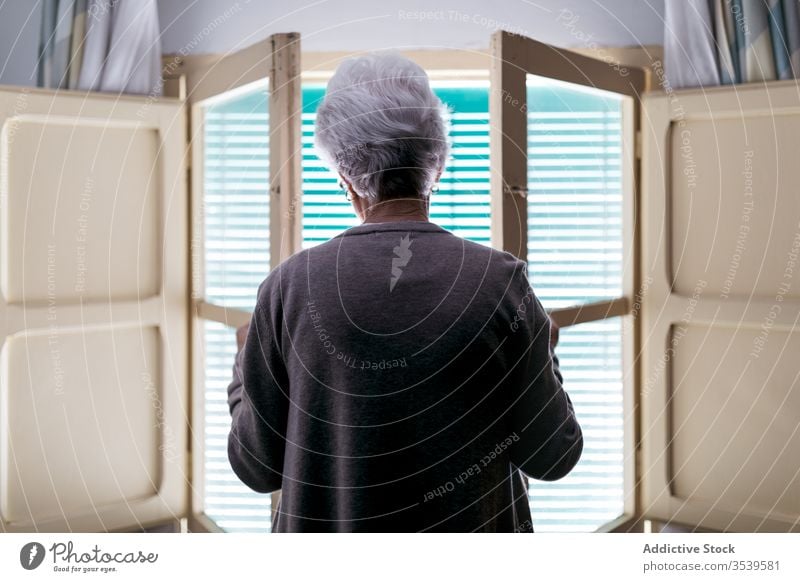 Gray haired woman standing in front of window at home senior lonely quarantine coronavirus shutter open alone old female solitude concept aged safety isolation