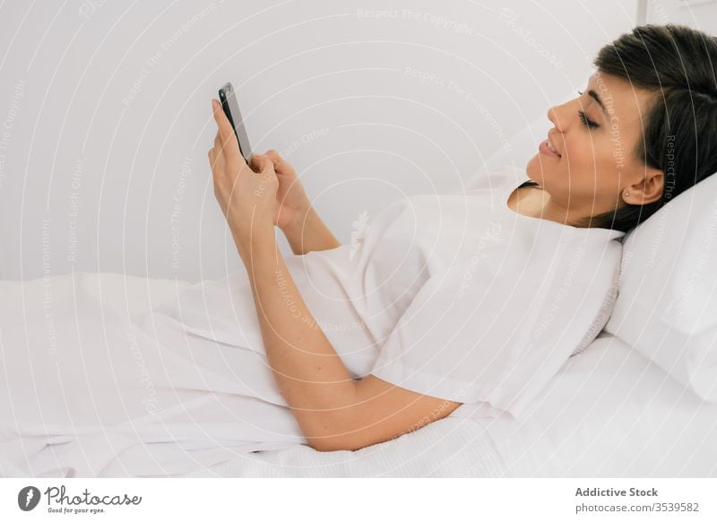Happy patient using smartphone in hospital ward woman lying down smile chair bed female modern robe cheerful white browsing clinic device texting gadget