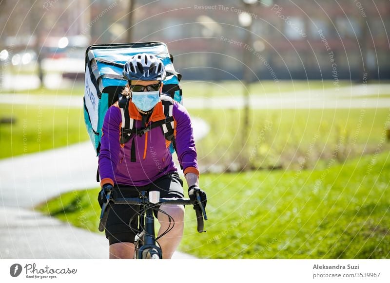Woman in helmet and protective mask with big food delivery backpack riding bike on the street. Sunny spring day in the city. Portrait of a female courier in a medical mask.