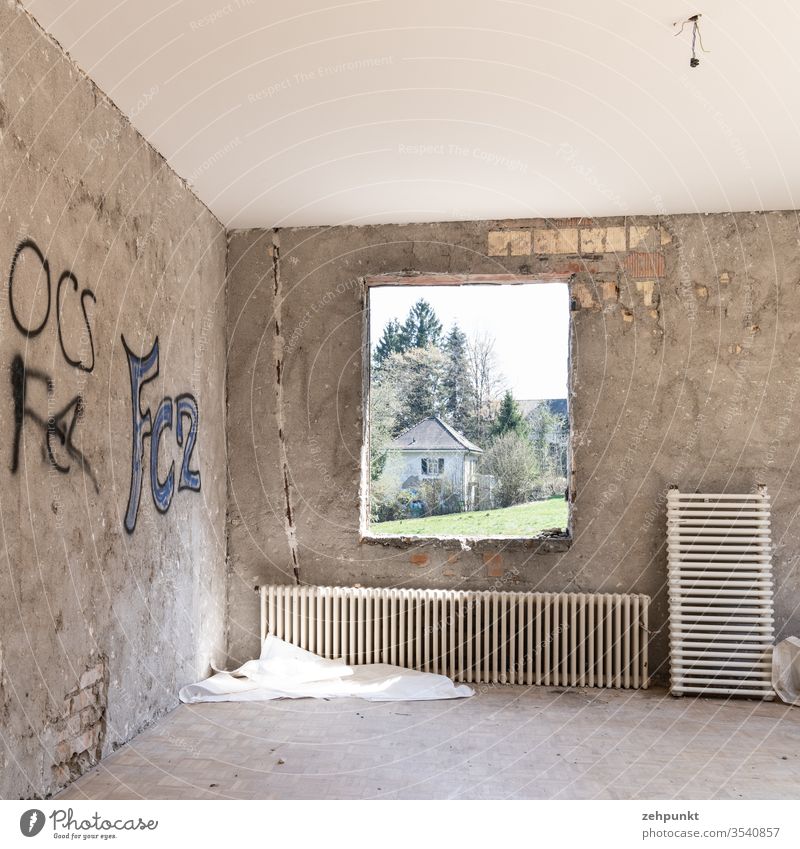 Room without furniture and without wall cladding, brickwork peeps out through plaster, two radiators leaning against the wall, graffiti on the walls, through frameless window opening views of meadow and next villa-like house