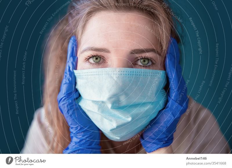 Young woman wearing protective gloves and face mask inside a home in quarantine looking bored and sad, for Covid-19 Coronavirus, with blue background medical
