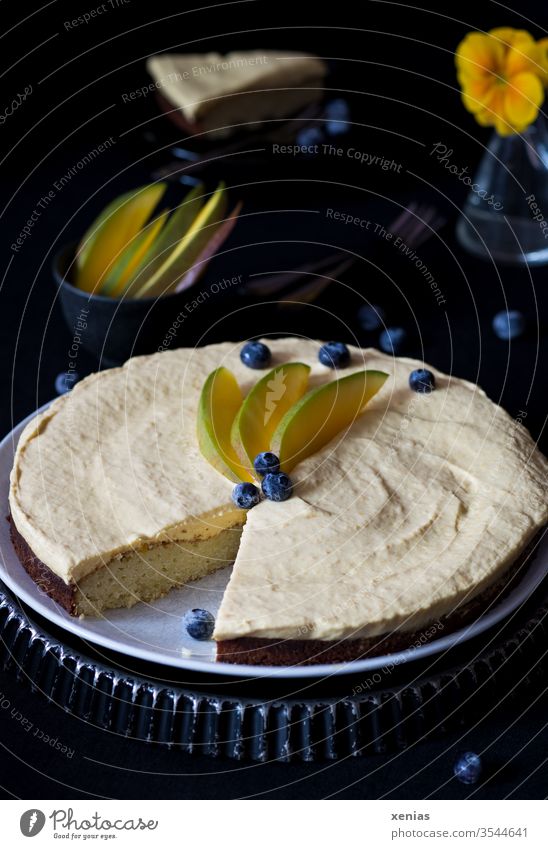 Sweet, round cake with white chocolate and mango cream, decorated with blueberries and mango slices, cake piece and vase in dark background Cake Mango