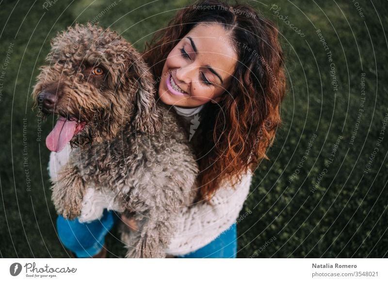 A beautiful woman is in the meadow with her dog. The owner is hugging her pet while looking at it with love. They are enjoying a day in the park. The pet is a Spanish water dog with brown fur.