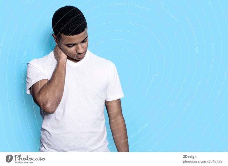 Closeup of young black man isolated on blue background in casual white t-shirt, touching neck with fingers looking down pensive and worried pain emotion male