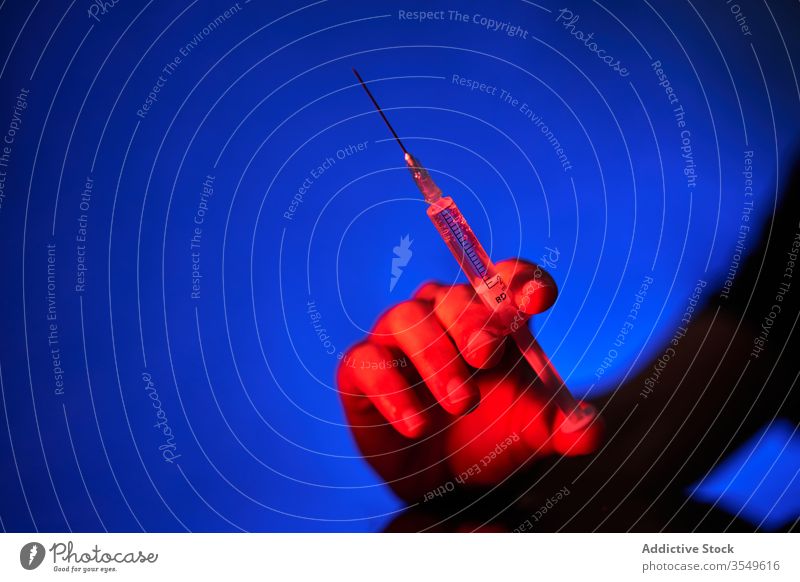 Crop person holding syringe with medication in red light on blue background vaccine cure injection hospital clinic covid 19 coronavirus infection disease