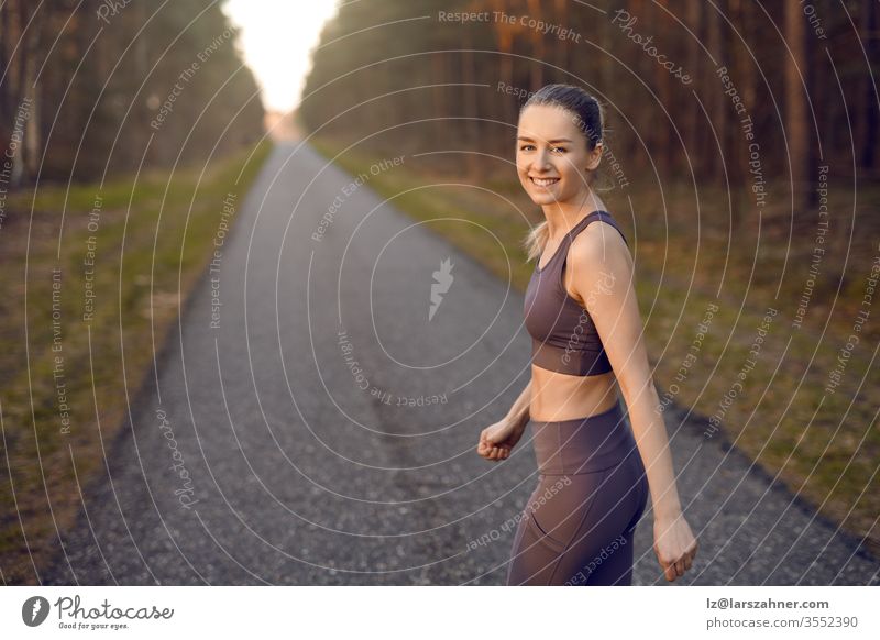 Fit athletic young woman out jogging at sunrise along a straight receding road through forest trees with glow of the sun at the end turning to smile at the camera in a healthy active lifestyle concept