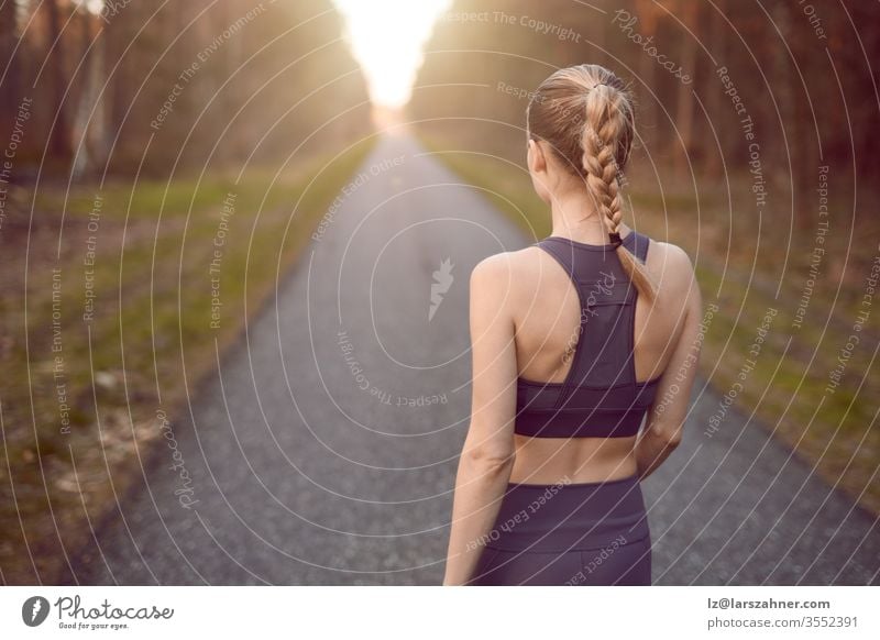 Sporty healthy young woman walking at sunrise along a rural road through a dense forest towards the glow of the sun at the end between the trees in a close up rear view