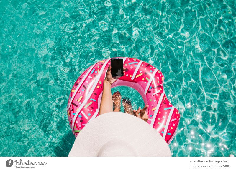 top view of a woman relaxing in the pool with pink donuts in hot sunny day. Summer holiday idyllic. Enjoying suntan Woman in bikini and a hat. Holidays and summer lifestyle. She is using mobile phone