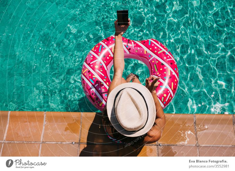 top view of a woman relaxing in the pool with pink donuts in hot sunny day. Summer holiday idyllic. Enjoying suntan Woman in bikini and a hat. Holidays and summer lifestyle. She is using mobile phone