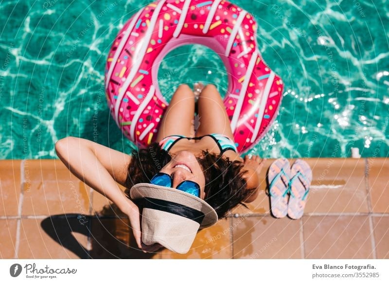 top view of a woman relaxing in the pool with pink donuts in hot sunny day. Summer holiday idyllic. Enjoying suntan Woman in bikini and a hat. Holidays and summer lifestyle
