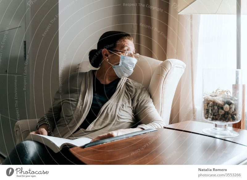 An older woman is sitting on the sofa with a book on her legs. She is looking through the window. She is sick and she is wearing a protective mask. She is protecting her family. Pandemic concept.