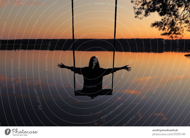 romantic young woman on a swing over lake at sunset. Young girl traveler sitting on the swing in beautiful nature, view on the lake sky water having fun playing