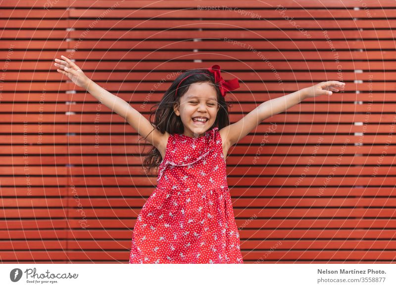 Happy little girl with arms wide open in front of a red background, wearing a red dress. summer fun cute kid funny happy hispanic child childhood young