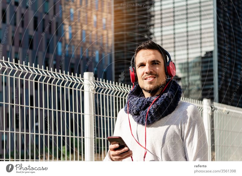 Young bearded male with headphones and holding smartphone while walking against skyscrapers in sunny day person man street outdoor earphones adult city