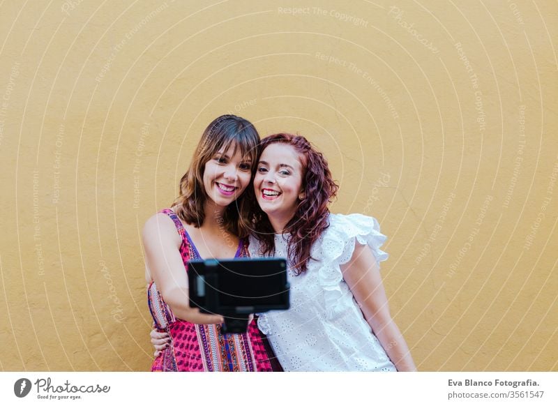 two friends or sisters taking a picture outdoors with mobile phone over yellow background. technology and lifestyle concept selfie city urban fun stick smiling