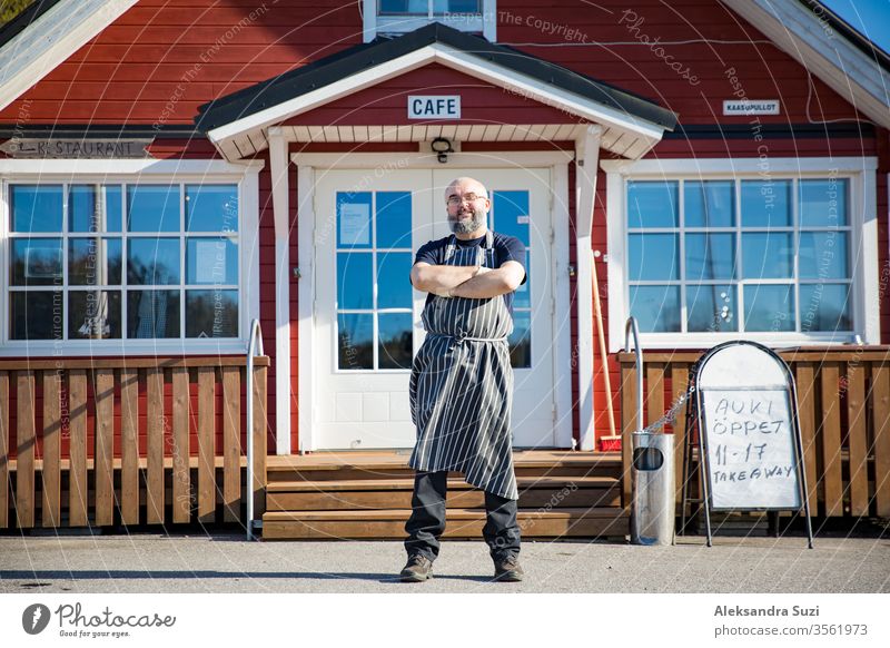 Portrait of a confident mature man with beard, restaurant employee wearing aprons standing in front of the restaurant. Small business concept. arms barista