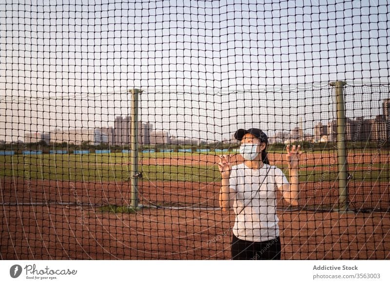 Young woman in protective mask standing behind net fence on sports ground coronavirus restriction prevent covid young sporty female asian ethnic safety