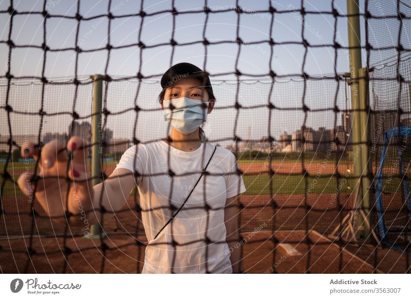 Young woman in protective mask standing behind net fence on sports ground coronavirus restriction prevent covid young sporty female asian ethnic safety