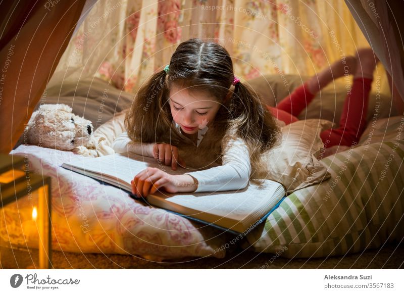 Cute little girl lying on pillows in homemade pink tent with flowers, read big book with interest and attention. Cozy stylish room. social distancing quarantine