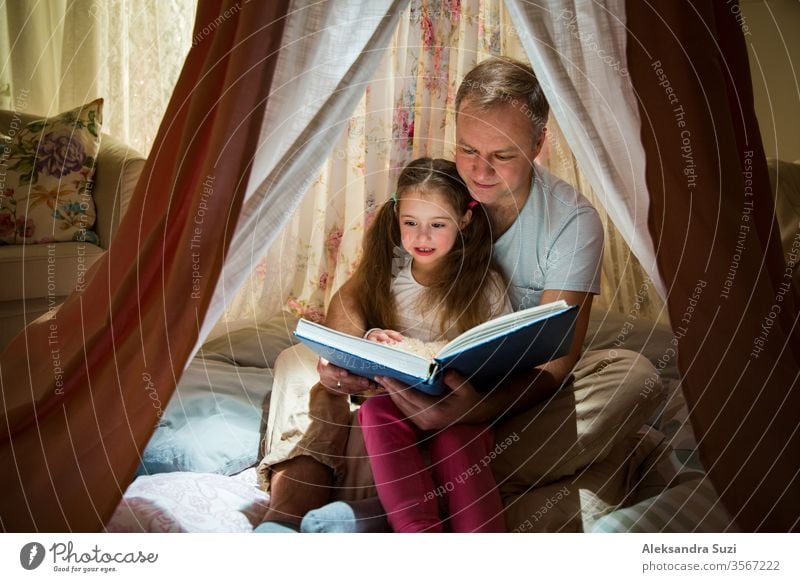 Family quality time. Father and daughter sit in homemade pink tent with flowers, read big book, look at each other, smile and laugh. Cozy stylish room. Family bonds concept