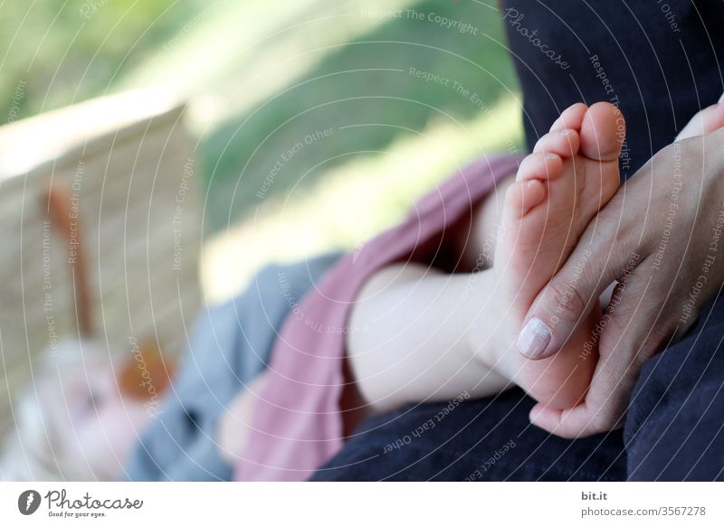 Hand of the mother holds tenderly, caring, lovingly the bare foot, of the sleeping child with pacifier, while falling asleep, outside at the sleeping place in nature. The toddler sleeps on a meadow in the garden and dreams peacefully.