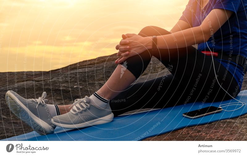 Woman sit and relax on yoga mat and listen to music via smartphone at stone beach by the sea . Woman outdoor workout. Fit girl wear smart band. Healthy, chill out lifestyle. Relax on summer vacations.