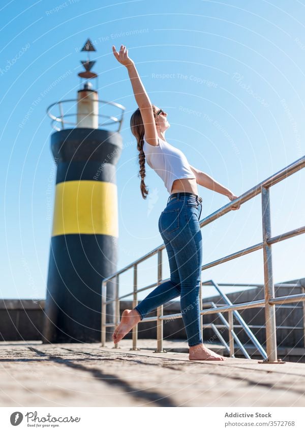 Content woman with raised arm at waterfront content dreamy embankment lighthouse enjoy seafront relax summer travel female cantabria spain casual barefoot stone