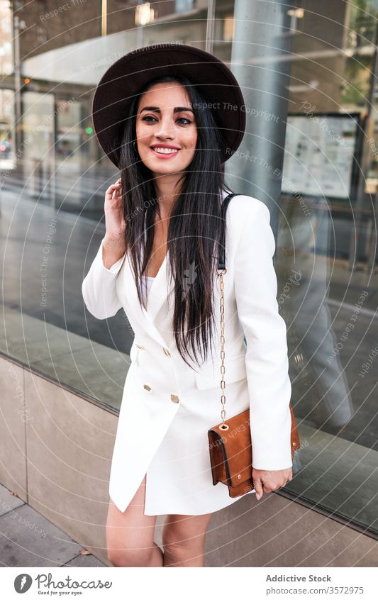 Fashionable young woman standing on city street style trendy fashion spring hat handbag season coat female positive modern confident building glass urban outfit