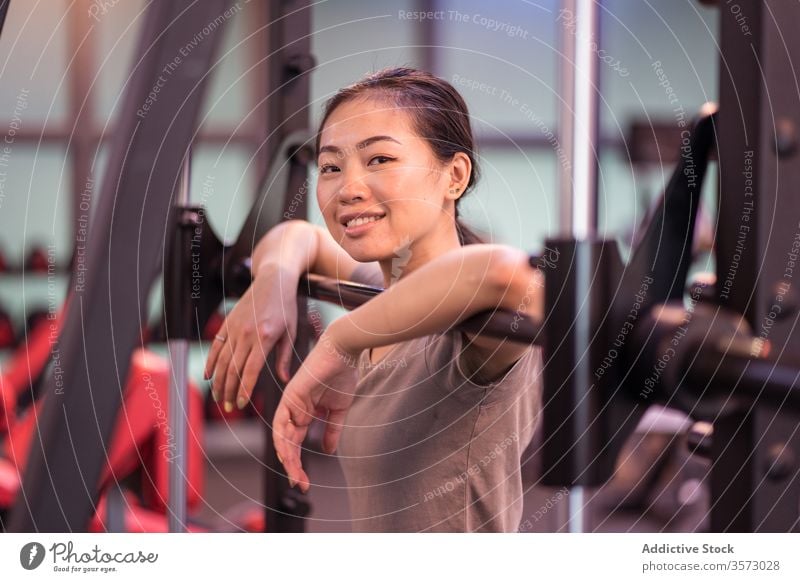 Positive Asian sportswoman leaning on barbell in modern gym and looking at camera rest workout machine content relax proud confident ethnic asian break fitness