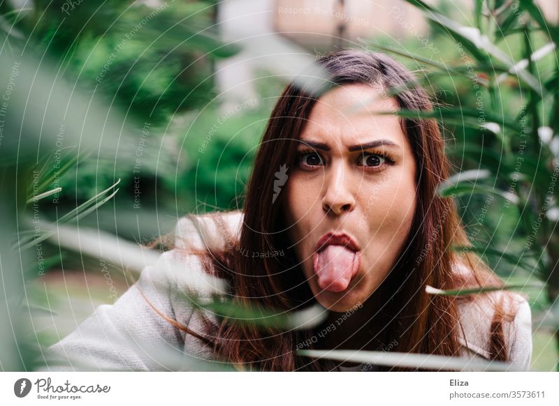 young woman looks evil behind plants in the garden and sticks out her tongue Evil stick out one's tongue Brash Sour Tongue Stick out Adults Human being 1