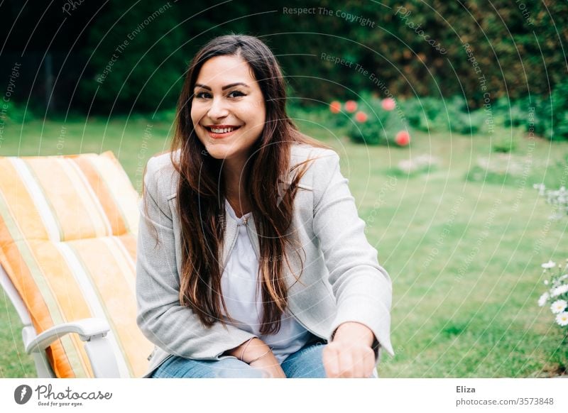 Brown-haired young pretty woman sitting on a deck chair in the garden and smiling into the camera Woman Garden out Sit Long-haired brown-haired brunette smile