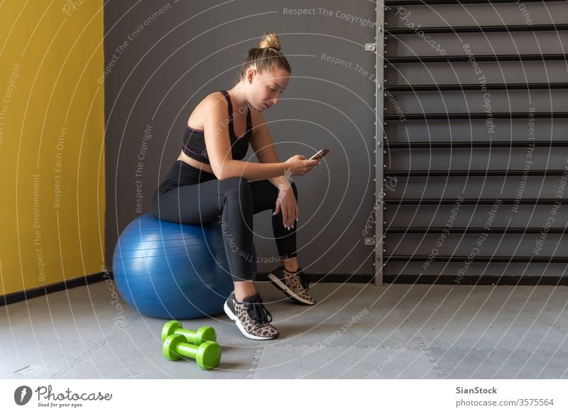 Sport woman sitting, resting and watching her mobile after workout or exercise in fitness gym. app phone sportive slim break dumbbells strength muscular strong