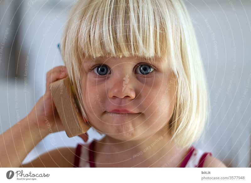 Cute, cute, blonde girl holds toy phone in hand and waits curiosity, eager for dial tone. Natural, cheerful child plays with wooden phone calling. Biological, ecological, sustainable, non-toxic children's toy.
