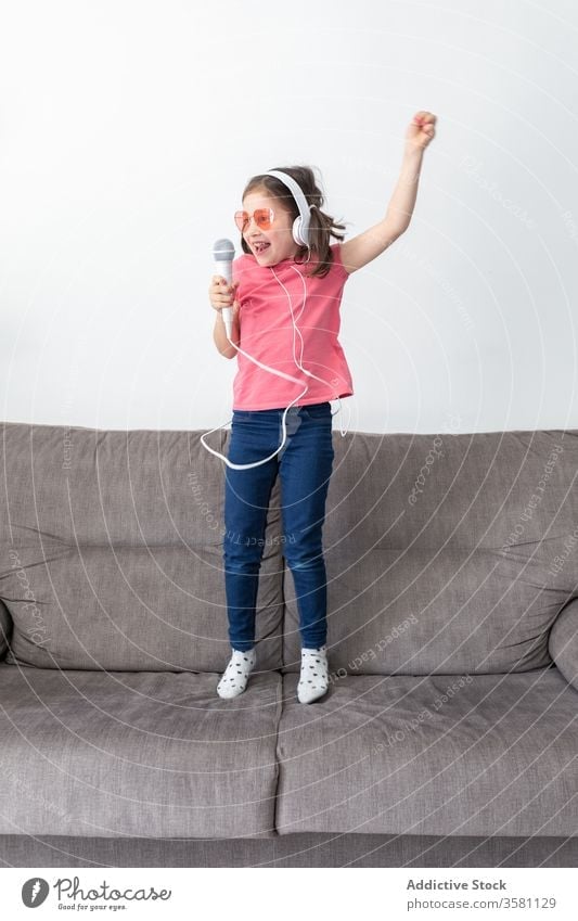 Happy preteen girl in headphones with mic singing while playing alone at home microphone having fun child jump sofa singer superstar happy music kid dance
