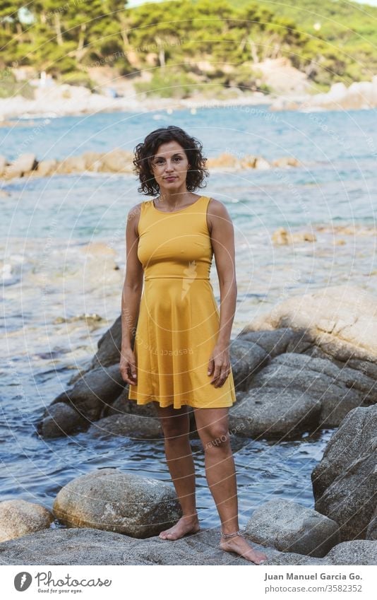 A beautiful female Latina wearing a yellow dress standing on a rock on the seashore woman girl beauty young pretty attractive people adult white natural person