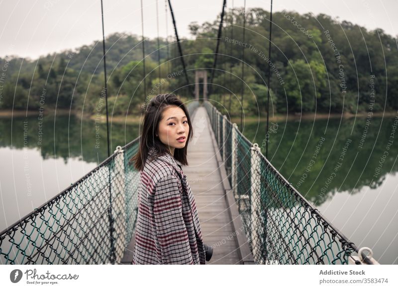 Calm Asian female traveler standing on suspension bridge over river tourist forest adventure tourism vacation picturesque peaceful journey serene reflection