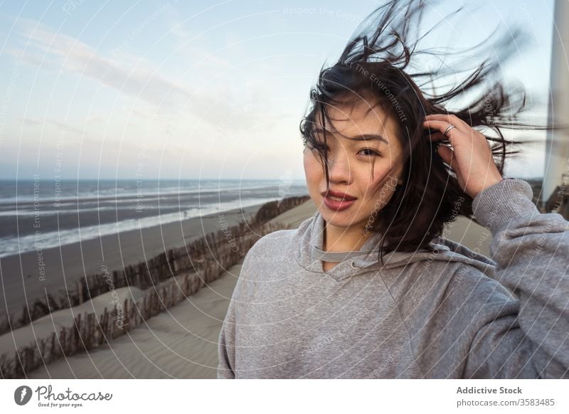 Young Asian woman standing on sea beach on windy weather skyline harmony sand touch hair coast thoughtful reflective calm coastline weekend recreation wistful