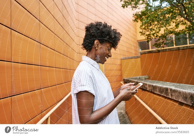 Happy black woman using smartphone on street against vivid exterior of modern building messaging pathway chill browsing use smile social media facade surfing