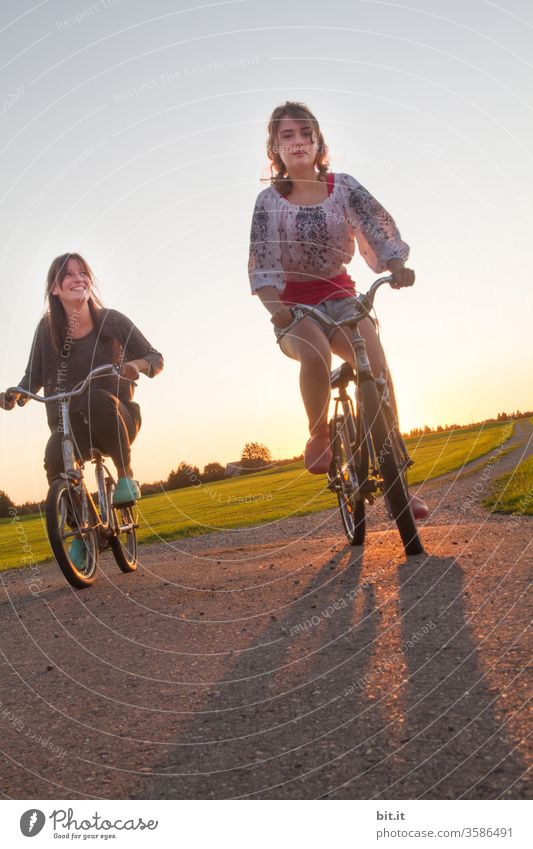 Two fashionable, pretty teenagers race together on old bikes in the evening sun. 2 feminine teenagers on a bike ride outside in the nature on the street. Sisters racing nostalgic bicycles in front of a green meadow on holiday.