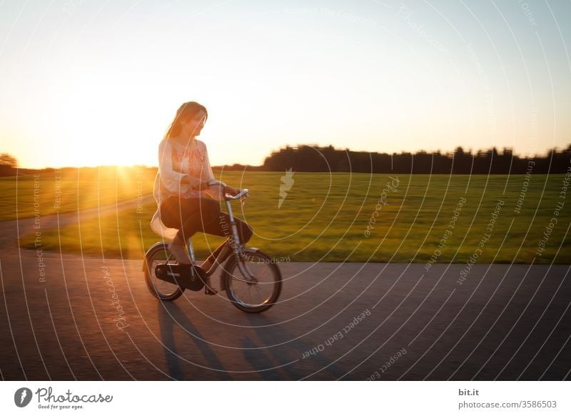 Long-haired, brunette teenagers, riding a small bicycle on the street in the evening light. Cycling in the twilight, passing green meadows and woods, the teenager, like a funny giant on the oversized, tiny dwarf bike.