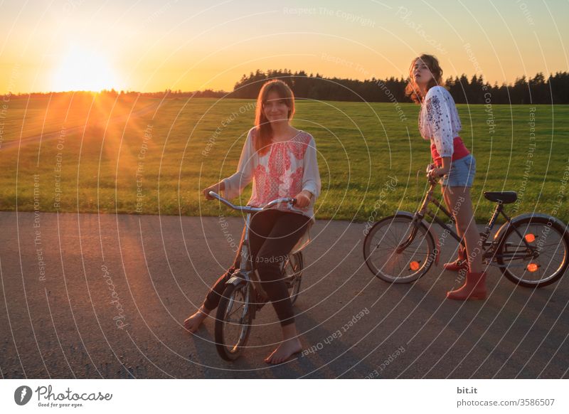Two long-haired, brunette teenagers, with fashionable, pretty blouses in hippie style, sit and stand on the street with old, nostalgic bicycles. 2 sisters pose in the evening sun with their bikes, in front of green meadows, in nature, on the street.