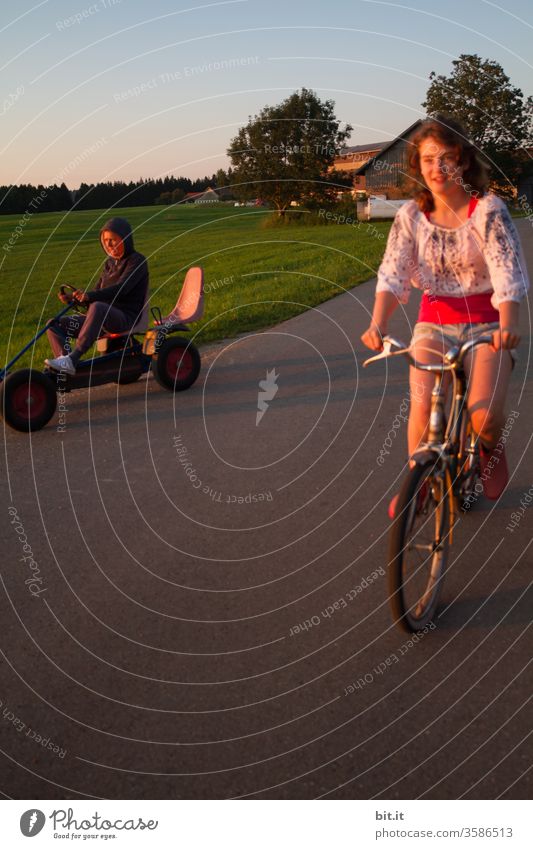 Mother and pretty daughter race together on old bike and Kettcar in the evening sun. Teenagers and adults on bike tours outside in the nature. Funny race with nostalgic vehicles in front of a green meadow on a farm holiday.