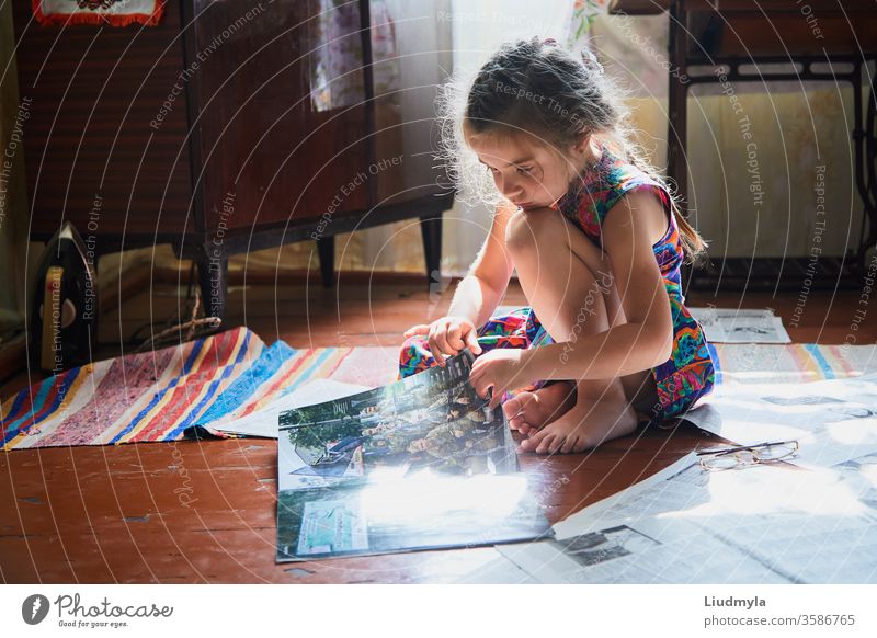 Little girl is sitting and looking into magazine on the floor inside the house. read newspaper curiosity children lifestyle contemporary experience knowledge