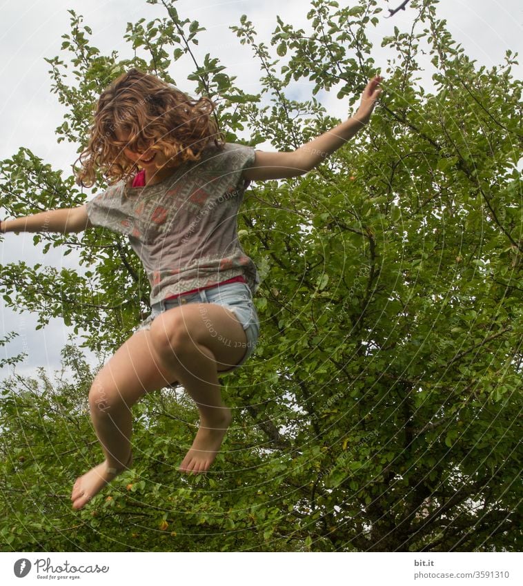 Curly, spirited joker jumps on trampoline in the garden. Girl flies, jumps, jumps lively, happy, wild, crazy up Sport, light-heartedness, exercise and fitness against stress, problems, fear at school and at home.