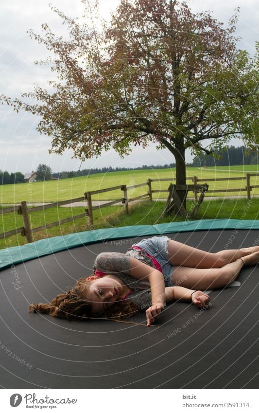 Tired, female teenager lies lazily on a trampoline in the garden and looks at his curly hair. Long-haired brunette teenager chills out on trampoline in nature, looking for split ends in strands of hair. Taking a break, resting, relaxing, recovering after sports.