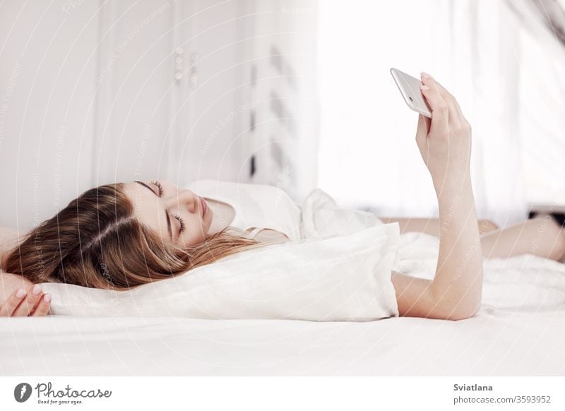Beautiful young girl looks at the phone while lying on the bed. A girl reads messages on her phone when she wakes up in the morning woman sleeping home pillow